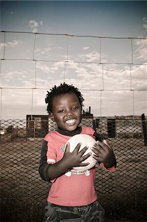 Young African schoolboy standing in a township, holding a soccer ball, smiling at camera Stock Photo - Premium Royalty-Free, Code: 6110-08968629