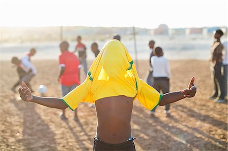 Boy celebrating with soccer jersey on his head in dirt field Stock Photo - Premium Royalty-Free, Code: 6113-06753765