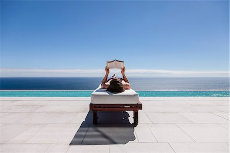 relaxing in poolside - Woman reading on lounge chair at poolside overlooking ocean Stock Photo - Premium Royalty-Free, Code: 6113-07543365
