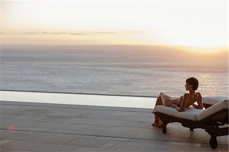 Woman in dress laying on lounge chair on patio overlooking ocean at sunset Stock Photo - Premium Royalty-Free, Code: 6113-07543388