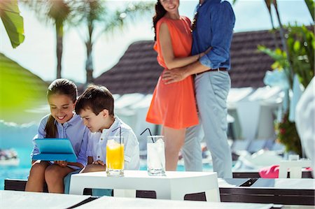 Brother and sister using digital tablet by resort swimming pool, parents embracing in background Photographie de stock - Premium Libres de Droits, Code: 6113-07808153