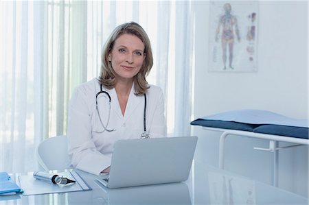 simsearch:6113-07905835,k - Portrait of smiling female doctor sitting at desk with laptop in office Foto de stock - Sin royalties Premium, Código: 6113-07808684