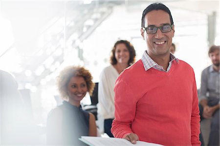 simsearch:6113-07808825,k - Portrait of smiling businessman wearing glasses and pink sweatshirt with team in background Stock Photo - Premium Royalty-Free, Code: 6113-07808899