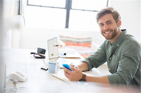 Portrait of young man sitting at desk with mobile phone and laptop Stock Photo - Premium Royalty-Free, Code: 6113-07992063