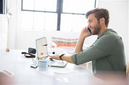 Portrait of young man sitting at desk with mobile phone and laptop Stock Photo - Premium Royalty-Free, Code: 6113-07992086