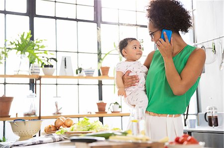 Mother holding her baby daughter while using mobile phone in kitchen Stock Photo - Premium Royalty-Free, Code: 6113-07992031