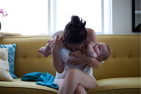Mother holding and kissing baby's belly, sitting on sofa by window Stock Photo - Premium Royalty-Free, Code: 6113-07906414