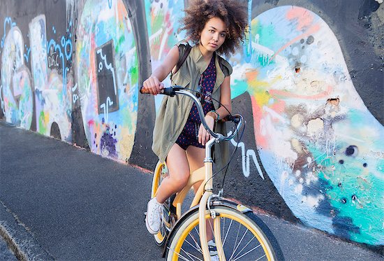 Portrait serious woman with afro on bicycle next to urban graffiti wall Stock Photo - Premium Royalty-Free, Image code: 6113-08171276