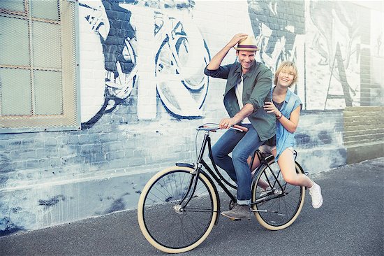 Portrait playful couple riding bicycle along urban graffiti wall Stock Photo - Premium Royalty-Free, Image code: 6113-08171290