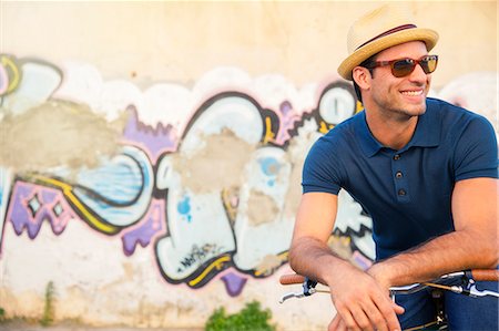 simsearch:6113-08171317,k - Smiling man in hat and sunglasses leaning on bicycle next to urban graffiti wall Stock Photo - Premium Royalty-Free, Code: 6113-08171317