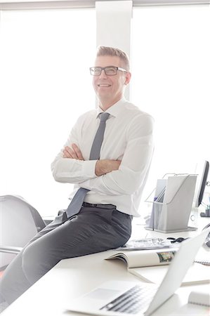 simsearch:6113-08805888,k - Portrait confident businessman with eyeglasses sitting on desk with arms crossed Photographie de stock - Premium Libres de Droits, Code: 6113-08171470