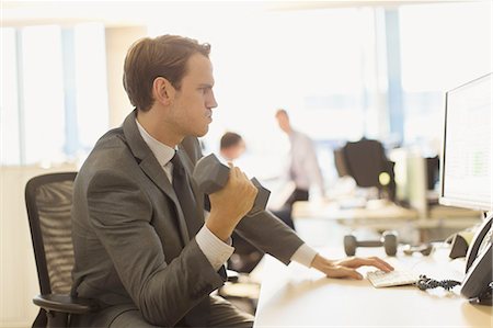 simsearch:6113-08171454,k - Businessman doing biceps curls with dumbbell at computer in office Fotografie stock - Premium Royalty-Free, Codice: 6113-08549945