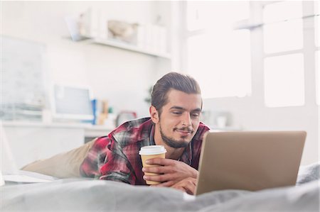 simsearch:6113-08568522,k - Young man drinking coffee at laptop on bed Fotografie stock - Premium Royalty-Free, Codice: 6113-08568554