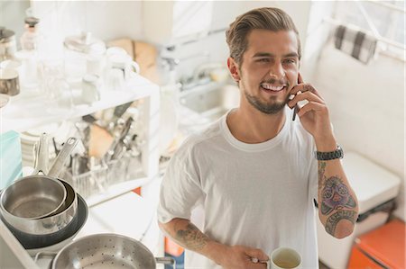 simsearch:649-09061281,k - Tattooed young man talking on cell phone and drinking coffee in apartment kitchen Stock Photo - Premium Royalty-Free, Code: 6113-08568528