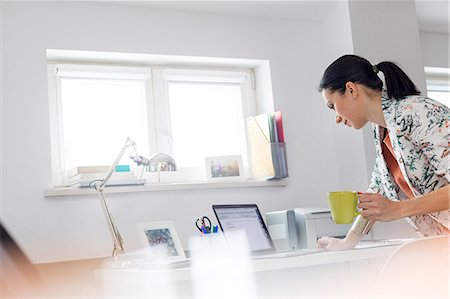 simsearch:6113-08743404,k - Businesswoman drinking coffee and working at laptop in office Stock Photo - Premium Royalty-Free, Code: 6113-08784435