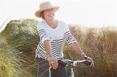 simsearch:6113-08985736,k - Smiling mature woman riding bicycle on sunny beach grass path Stock Photo - Premium Royalty-Free, Code: 6113-08985690