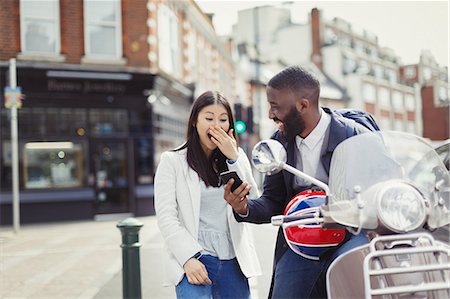 simsearch:6124-08926787,k - Young couple laughing, using cell phone at motor scooter on sunny urban street Fotografie stock - Premium Royalty-Free, Codice: 6113-09058514