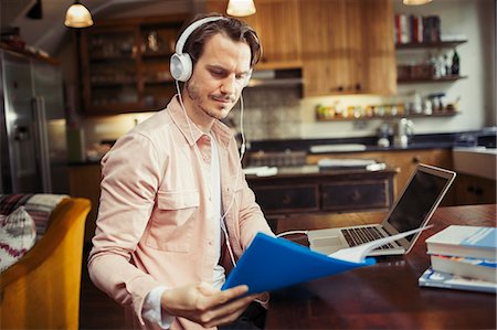 Man with headphones working at laptop, reading paperwork in kitchen Stock Photo - Premium Royalty-Free, Code: 6113-09059406