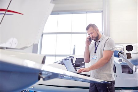 Male airplane mechanic talking on cell phone and working at laptop in hangar Stock Photo - Premium Royalty-Free, Code: 6113-09027789