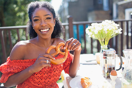 Portrait smiling, confident young woman with pretzel enjoying breakfast on sunny balcony Stock Photo - Premium Royalty-Free, Code: 6113-09220689