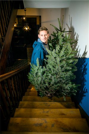 Young man carries Christmas tree upstairs Fotografie stock - Premium Royalty-Free, Codice: 6115-08104933