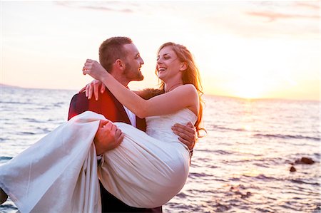 simsearch:6115-08239504,k - Groom carrying bride on pebble beach at sunset Stock Photo - Premium Royalty-Free, Code: 6115-08239495