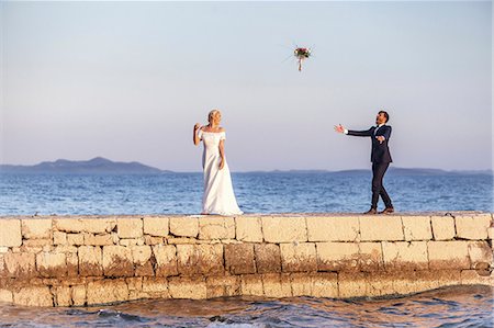 simsearch:6115-08239440,k - Bride and groom on pier throwing bouquet into air Foto de stock - Royalty Free Premium, Número: 6115-08239496