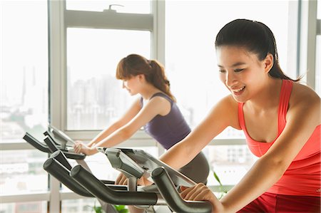 exercise indoors smile asian - Young women on stationary bikes exercising in the gym Stock Photo - Premium Royalty-Free, Code: 6116-06938630