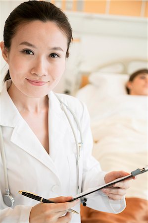 portrait doctor patient hospital - Portrait of smiling doctor holding a medical chart with patient lying in a hospital bed in the background Photographie de stock - Premium Libres de Droits, Code: 6116-07236092