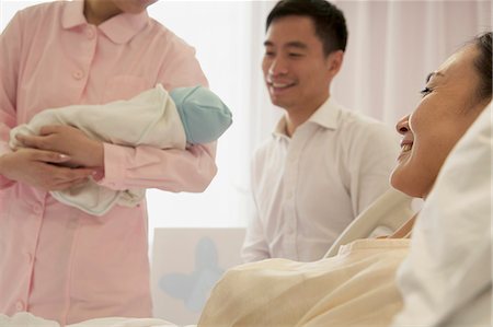 simsearch:6116-07236096,k - Nurse holding newborn baby in the hospital with mother lying on the bed and the father beside her Stock Photo - Premium Royalty-Free, Code: 6116-07236182