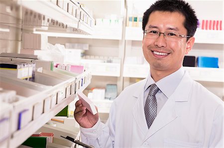 pharmacist helping - Pharmacist taking down and examining prescription medication in a pharmacy, looking at camera Stock Photo - Premium Royalty-Free, Code: 6116-07236102