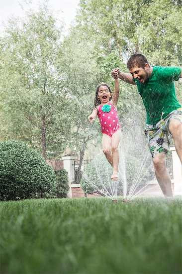 Father holding daughters hand while she jumps through the sprinkler in the garden Stock Photo - Premium Royalty-Free, Image code: 6116-07236226