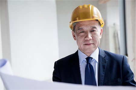 Smiling architect in a hardhat examining a blueprint in an office building Stock Photo - Premium Royalty-Free, Code: 6116-07236374