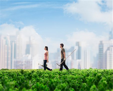 Two young people carrying gardening equipment walking across a green field with plants, cityscape in the background Stock Photo - Premium Royalty-Free, Code: 6116-07236483