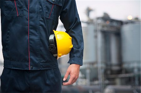 Midsection of young worker holding a yellow hardhat  outdoors with factory in the background Stock Photo - Premium Royalty-Free, Code: 6116-07236592