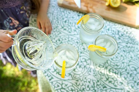 Making Lemonade. Overhead Shot Of Lemonade Glasses With A Fresh Slice Of Lemon In The Edge Of The Glass. A Child Pouring The Drink From A Jug. Foto de stock - Sin royalties Premium, Código: 6118-07121827