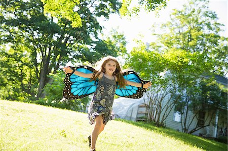 A Child Running Across The Lawn In Front Of A Farmhouse, Wearing Large Irridescent Blue Butterfly Wings And With Her Arms Outstretched. Stockbilder - Premium RF Lizenzfrei, Bildnummer: 6118-07121836