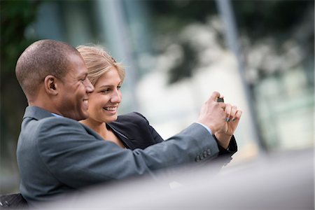 A Young Blonde Woman And A Man On A New York City Street. Wearing Business Clothes. Standing Side By Side Taking A Selfy Photograph With A Smart Phone. Photographie de stock - Premium Libres de Droits, Code: 6118-07121956