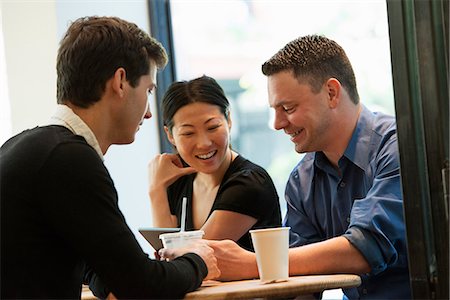 A Group Of People Sitting Around A Table In A Coffee Shop. Looking At The Screen Of A Digital Tablet. Two Men And A Woman. Stock Photo - Premium Royalty-Free, Code: 6118-07122296