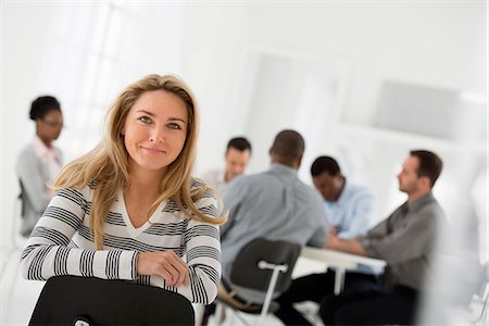 simsearch:6118-07122641,k - Office Interior. A Woman In A Striped Shirt Seated Astride A Chair Looking Away From A Group Seated At A Table. Stock Photo - Premium Royalty-Free, Code: 6118-07122690