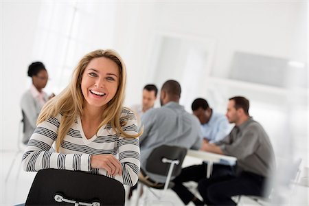 Office Interior. A Woman In A Striped Shirt Seated Astride A Chair Looking Away From A Group Seated At A Table. Foto de stock - Sin royalties Premium, Código: 6118-07122691