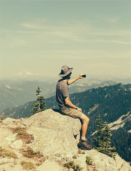 A hiker on a mountain summit, holding a smart phone, at the top of Surprise Mountain, in the Alpine Lakes Wilderness, in Mount Baker-Snoqualmie National Forest. Stock Photo - Premium Royalty-Free, Image code: 6118-07202945