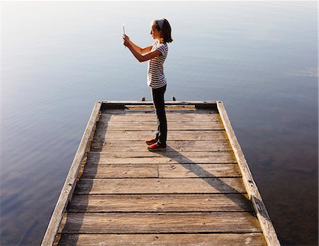 A young girl holding a digital tablet in front of her, standing on a wooden dock over the water. Stock Photo - Premium Royalty-Free, Code: 6118-07202935