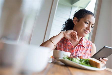 A young woman reading from the screen of a digital tablet, seated at a table. Coffee and a sandwich. Stock Photo - Premium Royalty-Free, Code: 6118-07203657