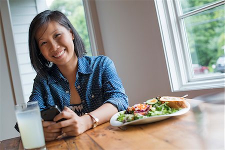 simsearch:6118-07203660,k - A young woman using a smart phone, seated at a table. Coffee and a sandwich. Fotografie stock - Premium Royalty-Free, Codice: 6118-07203658