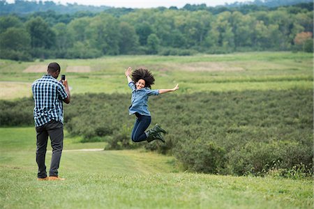 simsearch:6118-07203656,k - A man taking a photograph of a woman leaping in the air, jumping for joy with her arms outstretched. Fotografie stock - Premium Royalty-Free, Codice: 6118-07203676