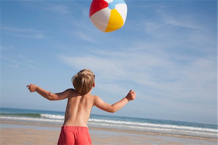 A boy in swimming trunks on the beach, with a large beach ball in the air above him. Stock Photo - Premium Royalty-Free, Code: 6118-07203841