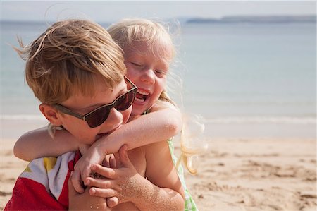 A brother and sister play fighting on the beach. A boy in sunglasses and a younger girl with her arms around his neck. Stock Photo - Premium Royalty-Free, Code: 6118-07203849