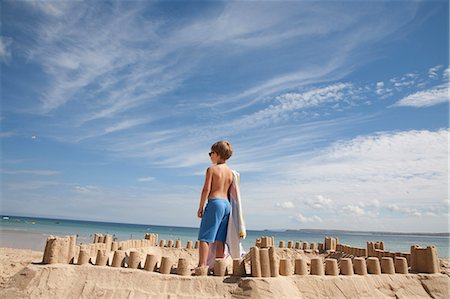 A boy standing beside a sandcastle, on top of a mound of sand. Beach. Foto de stock - Royalty Free Premium, Número: 6118-07203856