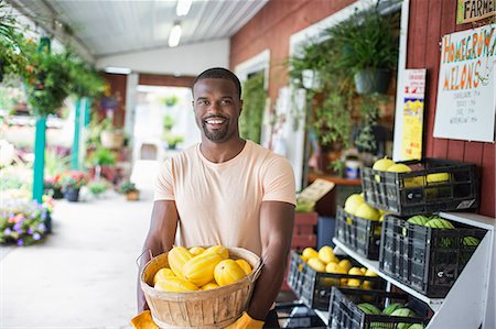 Working on an organic farm. A man carrying a large basket of yellow squash vegetables. Displays of fresh produce for sale. Foto de stock - Royalty Free Premium, Número: 6118-07203880